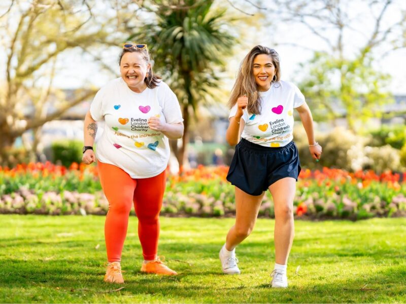 Two Evelina Children's Charity supporters participating in 'Step for Evelina' event. They smiling and walking amongst grass with flowers and trees in the background.