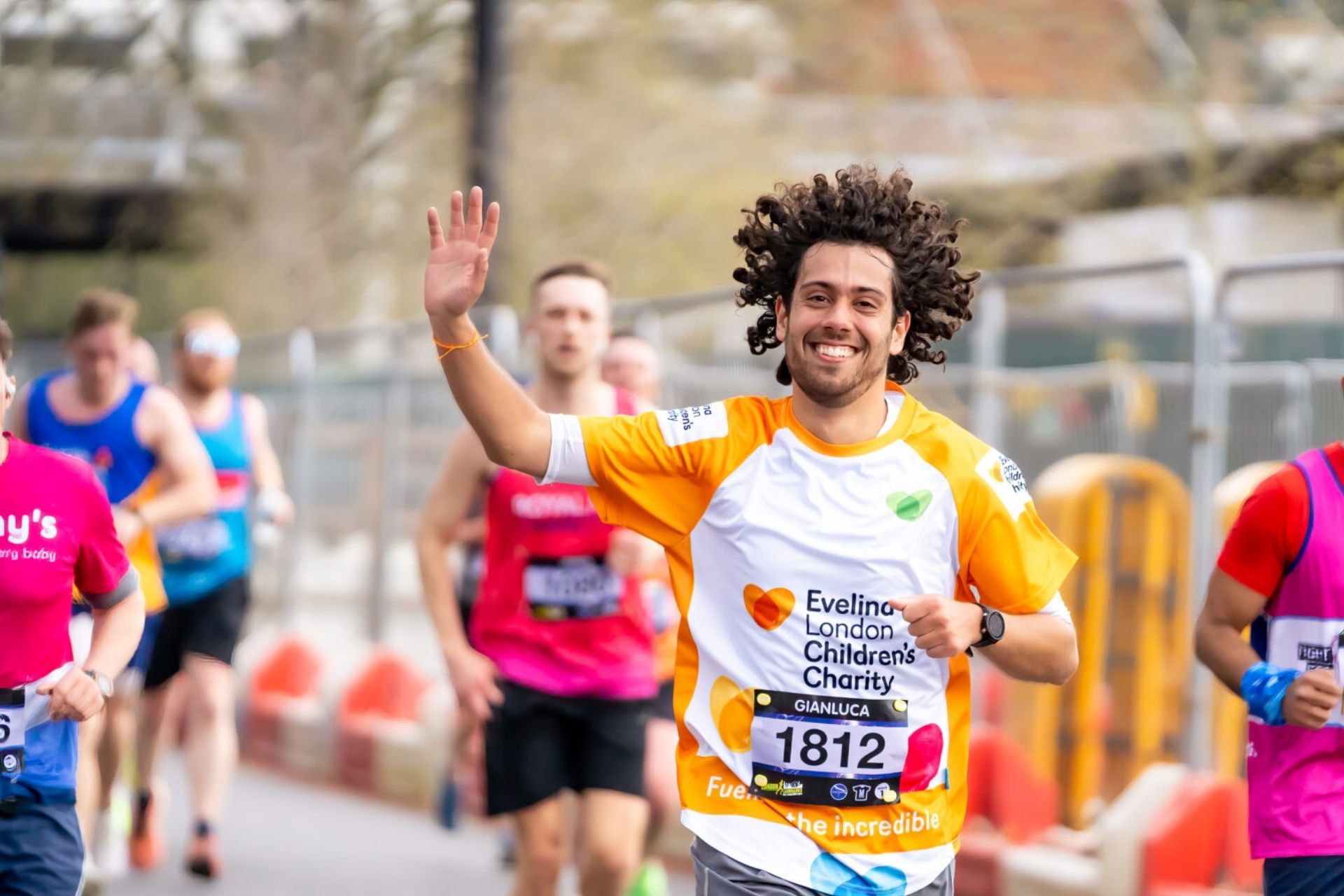 Evelina London Charity Supporter running in a marathon alongside other runners. He appears to be smiling and waving, wearing an orange Evelina London Children's Charity T-shirt