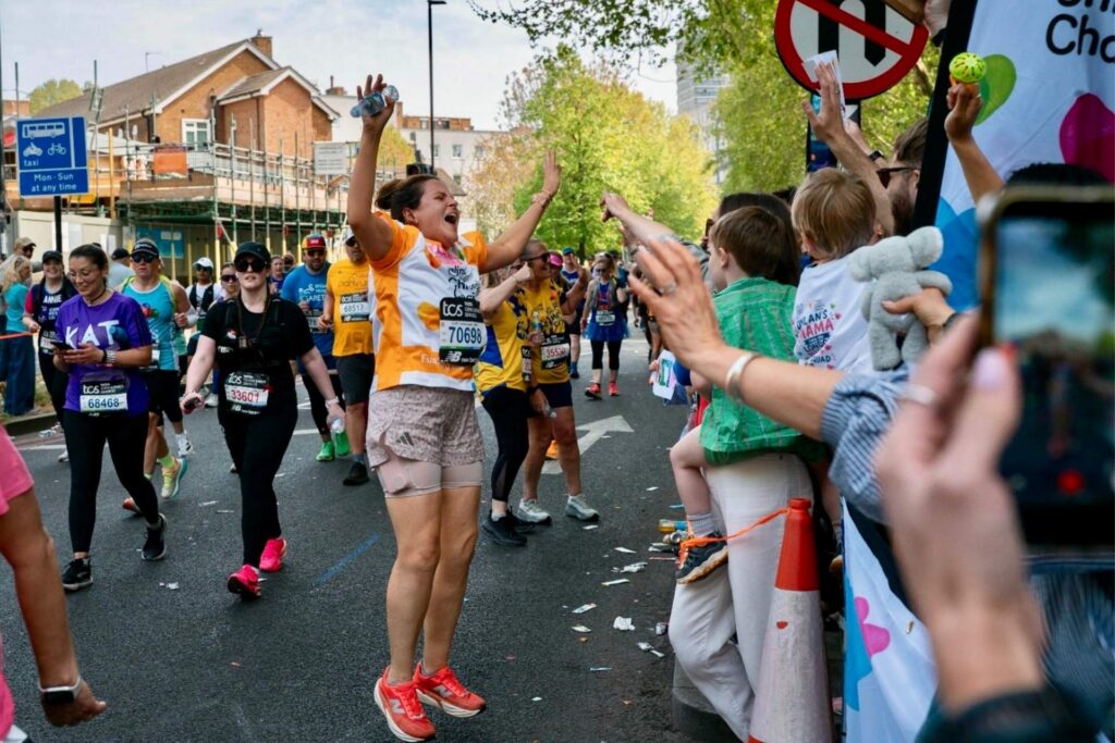 Anita is jumping in the air with her arms up high, in front of her is her cheer squad cheering her on at London Marathon. Anita is wearing an orange Evelina London Children