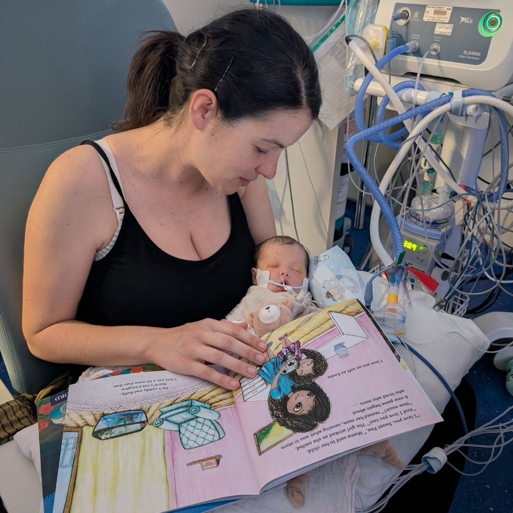 Becky is sat in a blue chair, reading a story book to her daughter Ada who is laid in her arms. Beside them is machinery and they are in Evelina London Children