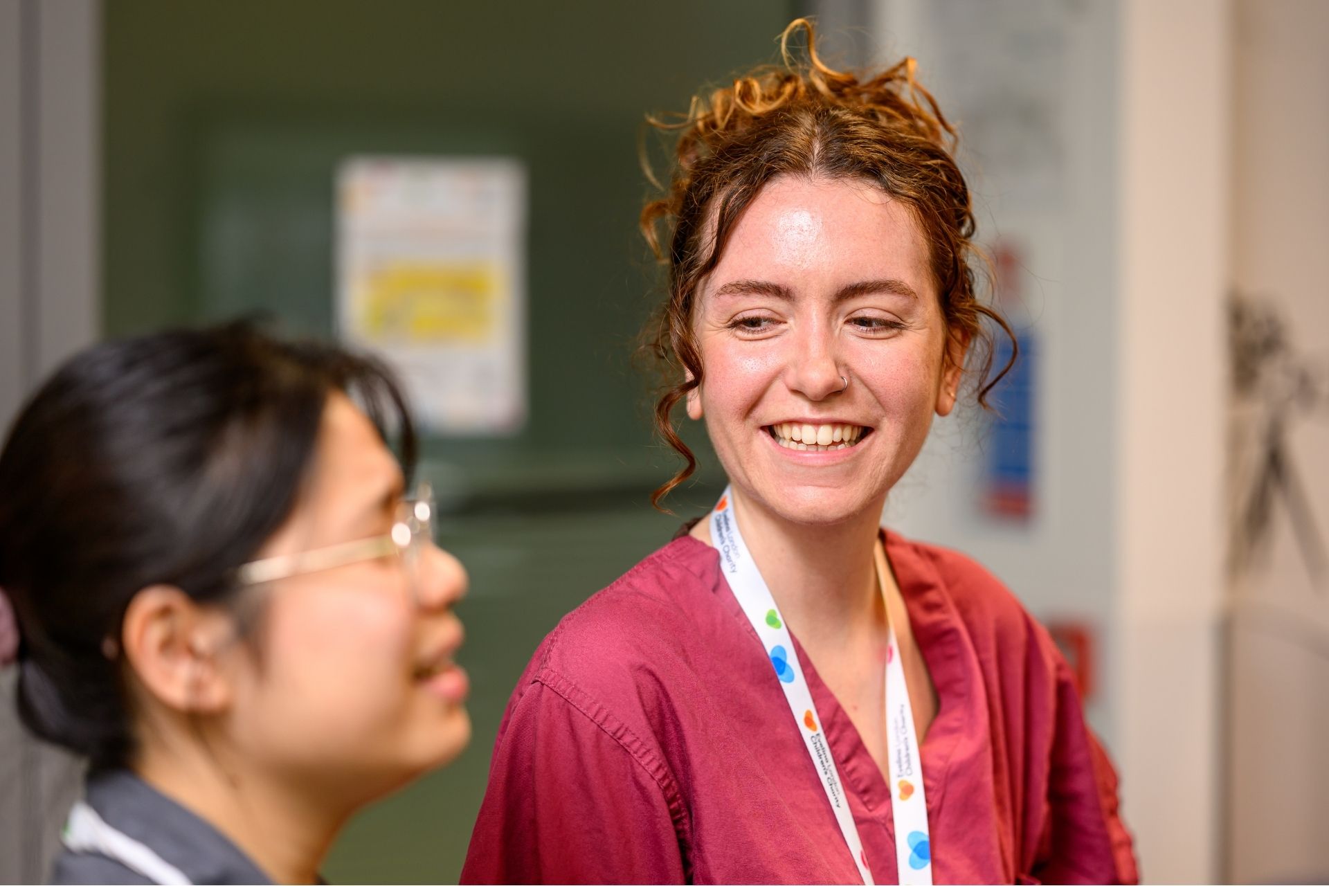 Two staff members at Evelina London Children's Hospital, one on the left with dark brown hair is slightly blurred, in focus on the right appears to be smiling, wearing a dark pink uniform, white lanyard reads 'Evelina London Children's Charity'.
