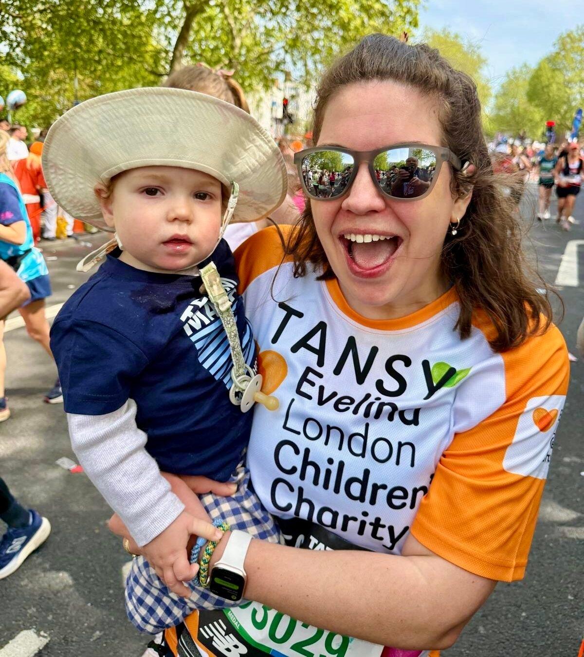Tansy and Atlas at London Marathon event 2026. Tansy wearing sunglasses and appears to be smiling in an orange Evelina Children's Charity t shirt holding in her arms her son, Atlas who is wearing a navy blue top, with a long white sleeve top underneath and a cream coloured hat.