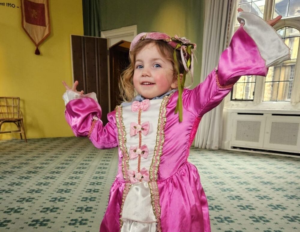 Alana is in a pink dress, arms up in the air smiling, in the background is a tiled floor with yellow walls.