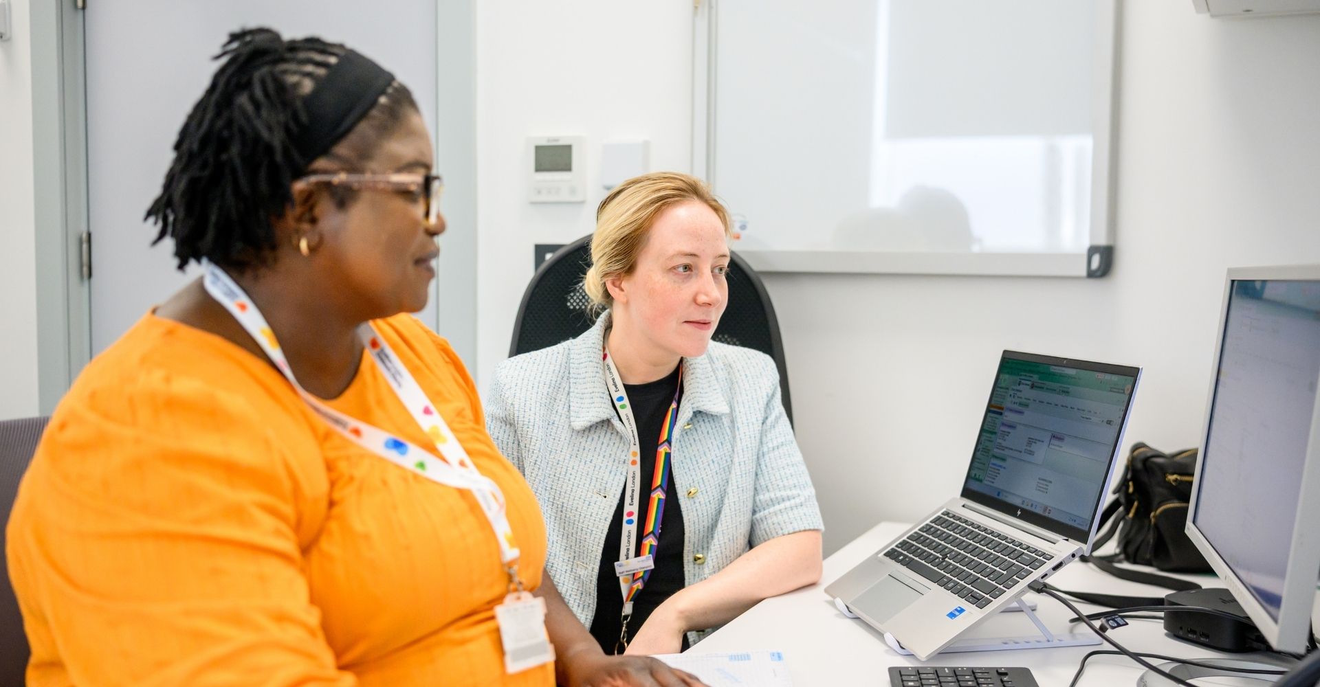Two members of staff from Evelina London Children's Hospital. They're sat down looking at a computer monitor together, the person on the left is wearing an orange top and next to them they're in a patterned top.
