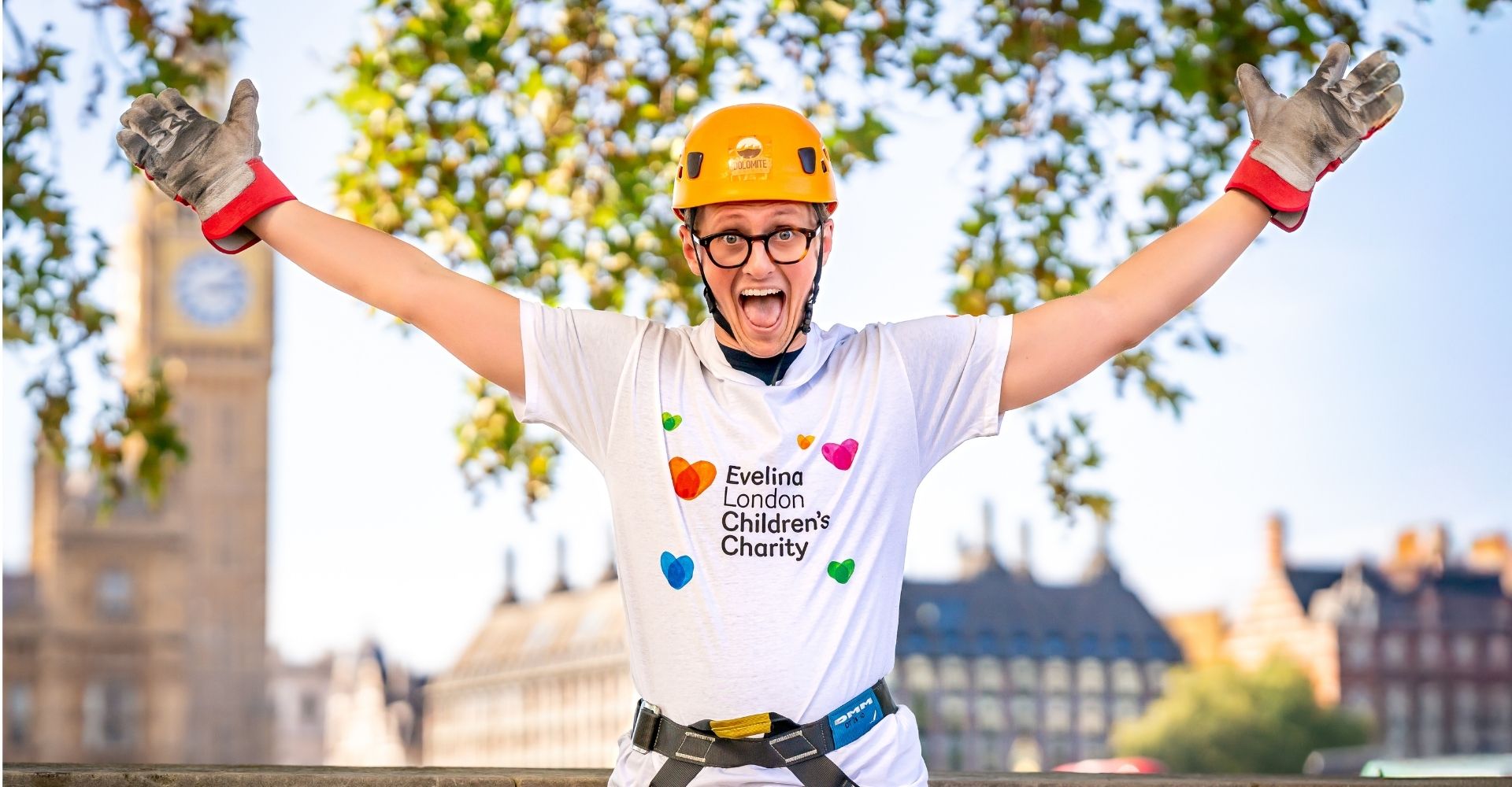 Evelina London Children's Charity Supporter, excitedly raising their arms up, wearing an orange helmet and a white charity t shirt with the logo and heart motifs.