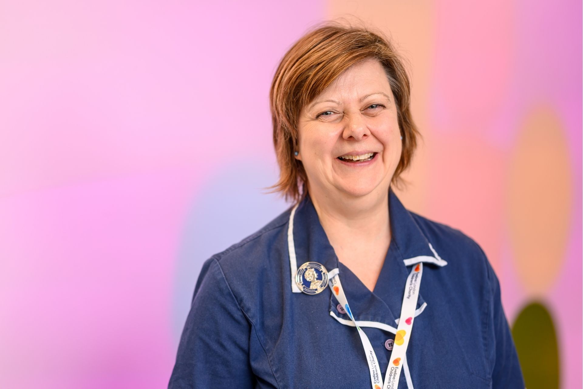 An Evelina London Children's Hospital member of staff appears to be smiling in a blue uniform, in the background it is a light pink and orange.