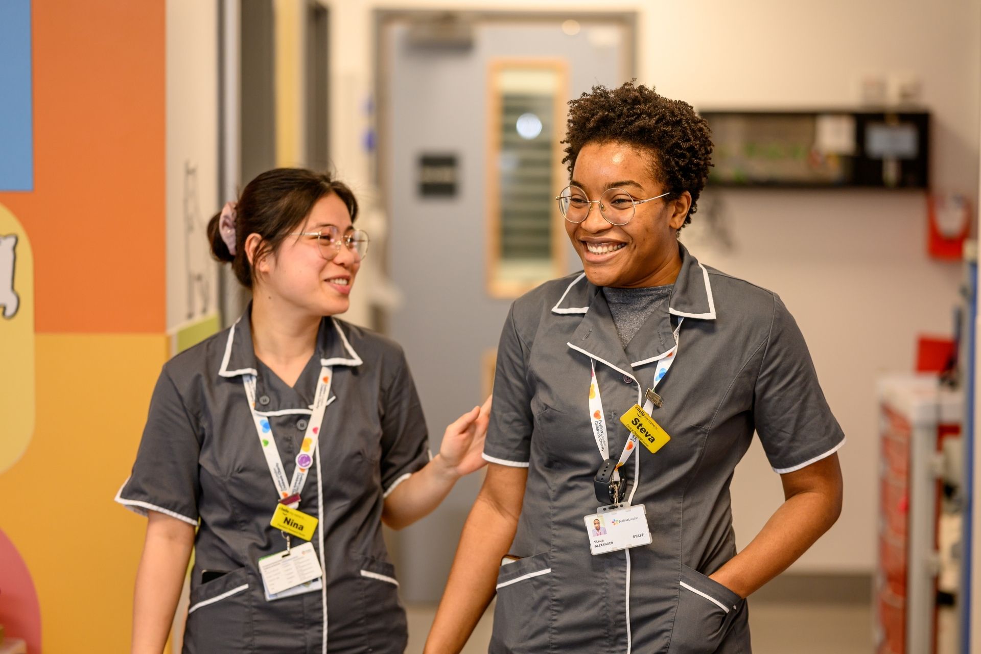 Two Evelina London members in grey uniform with white piping on their collars, they appearing to be smiling.