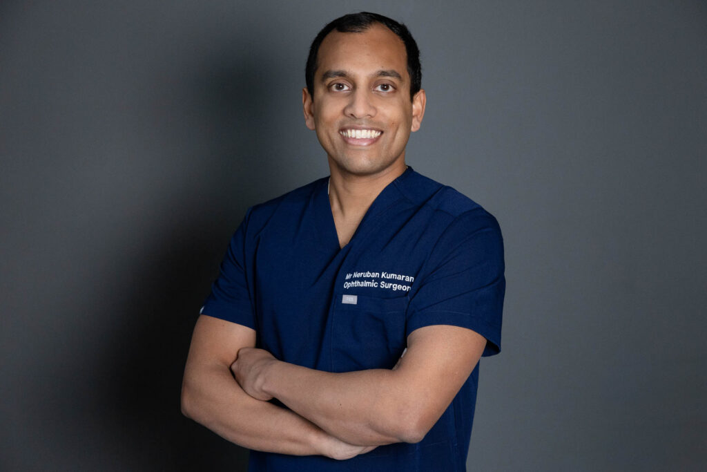 Dr Neruban Kumaran with his arms folded, he appears to be be smiling and is wearing a dark navy blue uniform. The wall in the background is a dark grey.