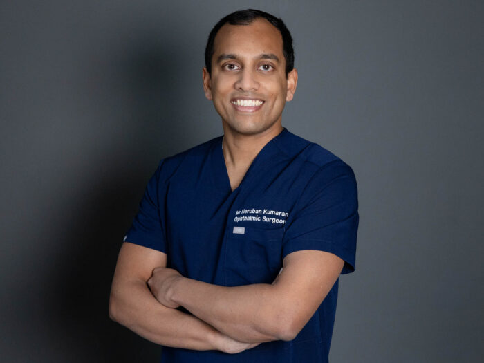 Dr Neruban Kumaran with his arms folded, he appears to be be smiling and is wearing a dark navy blue uniform. The wall in the background is a dark grey.