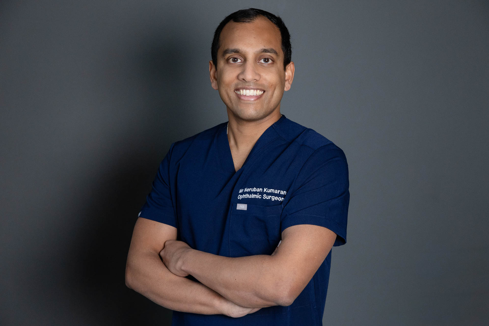 Dr Neruban Kumaran with his arms folded, he appears to be be smiling and is wearing a dark navy blue uniform. The wall in the background is a dark grey.