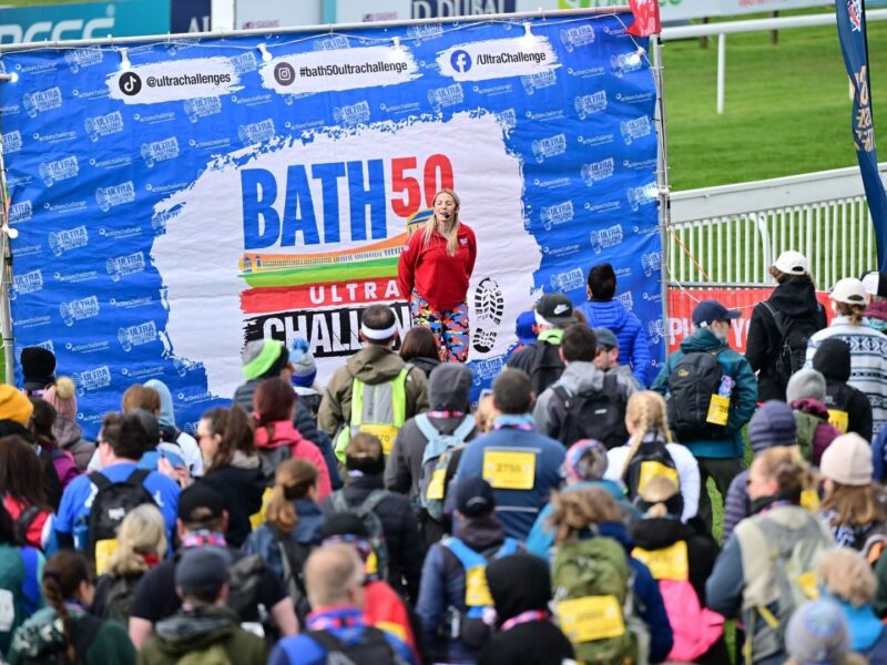 Challengers of the Bath 50 Ultra are being briefed at the start. They're on a stage and in the background it reads 'Bath 50 Ultra Challenge' in various colours and different size text repeatedly.