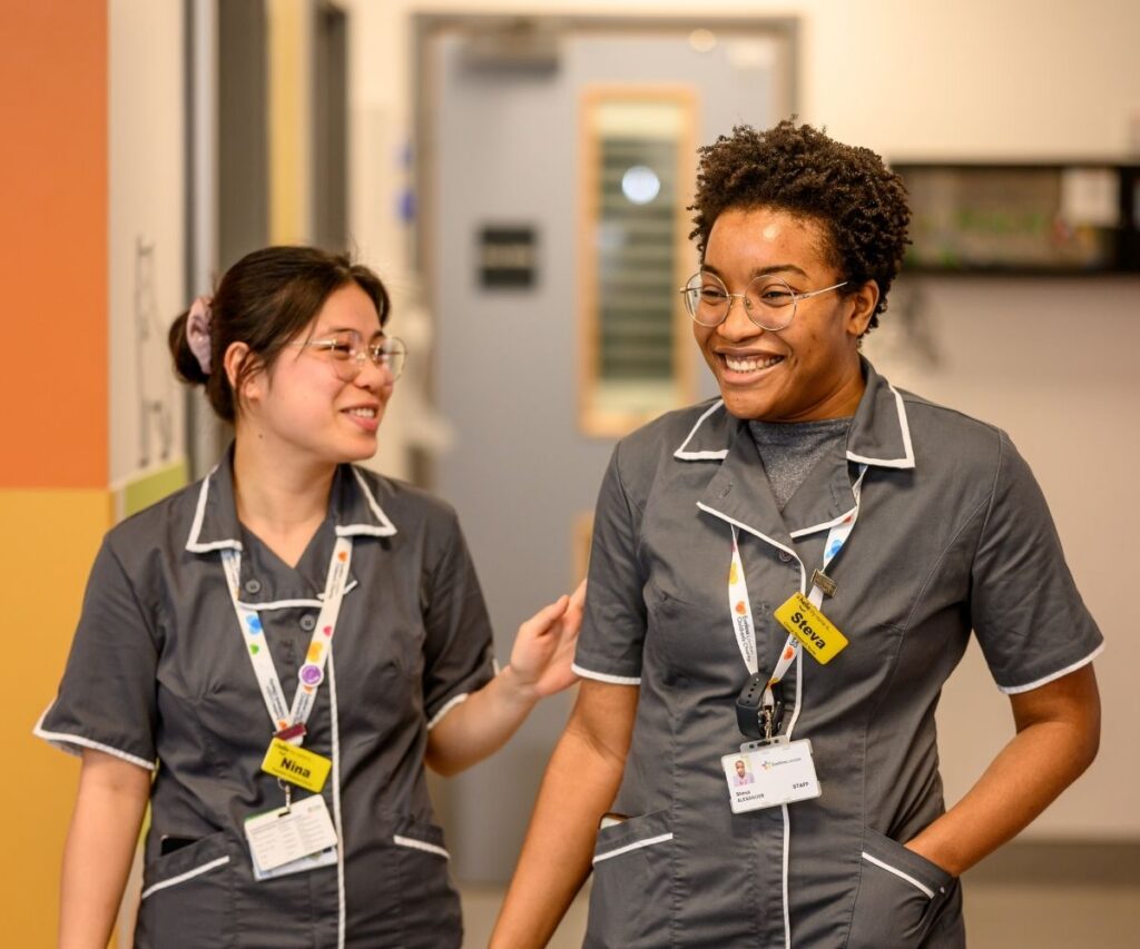 Two Evelina London members in grey uniform with white piping on their collars, they appearing to be smiling.